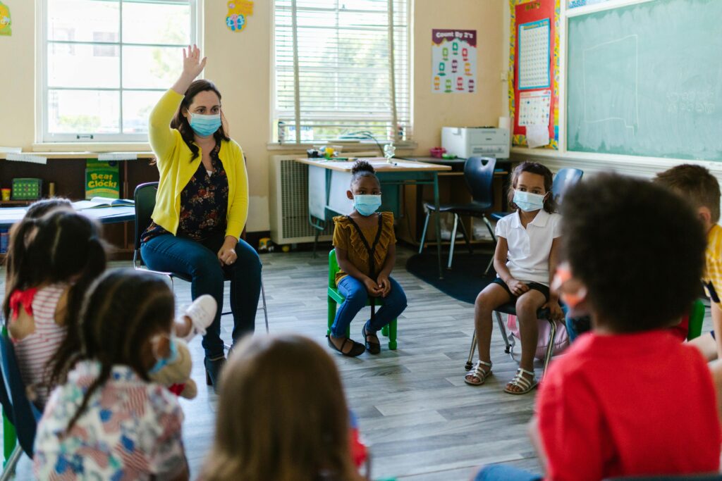 Teacher and children in a masked preschool setting engaged in learning activities indoors.